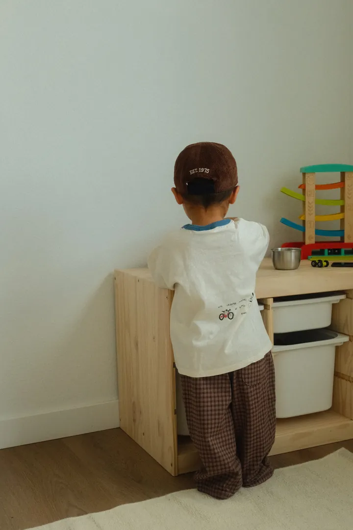 A toddler seen from behind in a brown corduroy cap, cream tee with a blue collar and brown gingham trousers, playing at a wooden toy shelf
