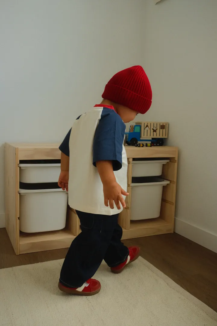 A toddler in a red ribbed beanie, navy and cream raglan tee, dark jeans and red sneakers, walking past a wooden toy shelf